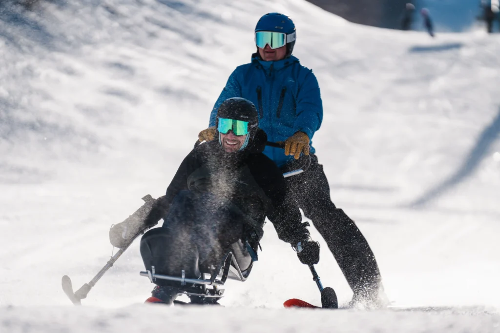 Two individuals are skiing down a snow-covered slope, one assisting another in a specialized sled, creating a cloud of snow dust.