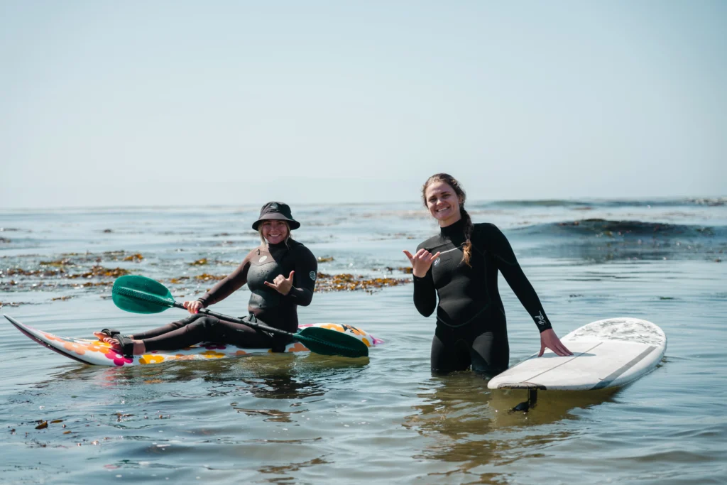 Two individuals in wetsuits enjoying the water; one is sitting on a colorful kayak while the other stands beside a surfboard.