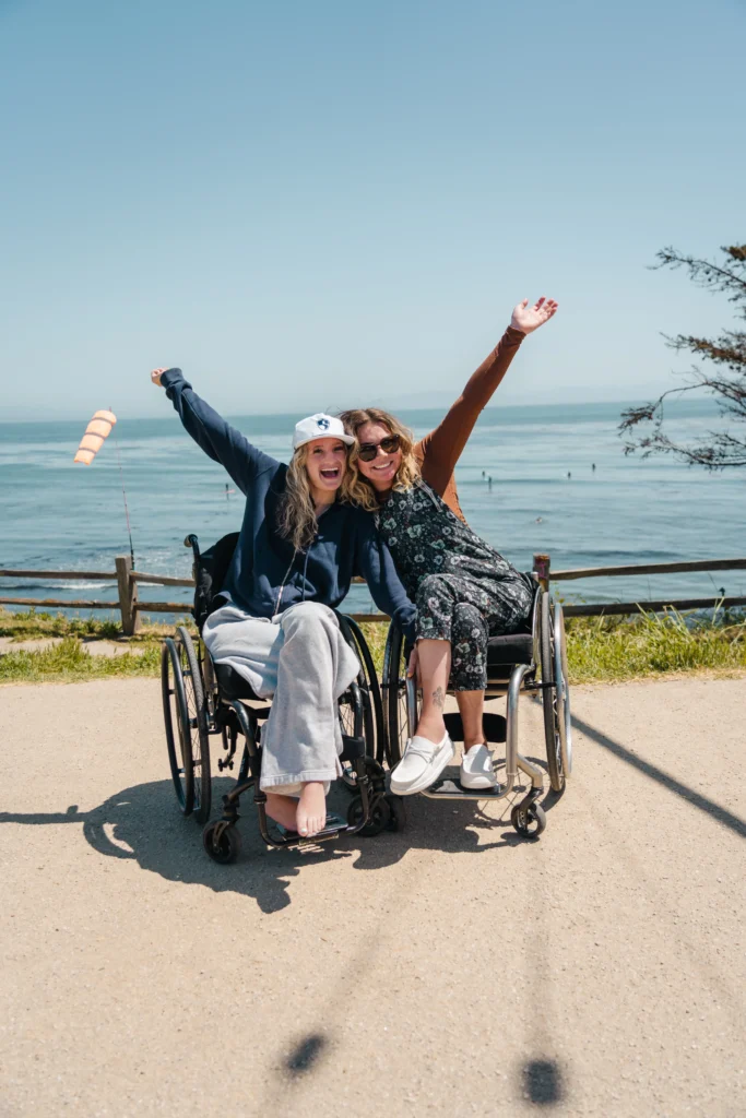 A group of wheelchair users and standing individuals poses together on a sunny beach, smiling and making hand gestures, with the ocean in the background.