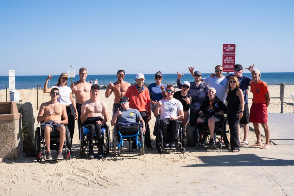 A group of wheelchair users and standing individuals poses together on a sunny beach, smiling and making hand gestures, with the ocean in the background.