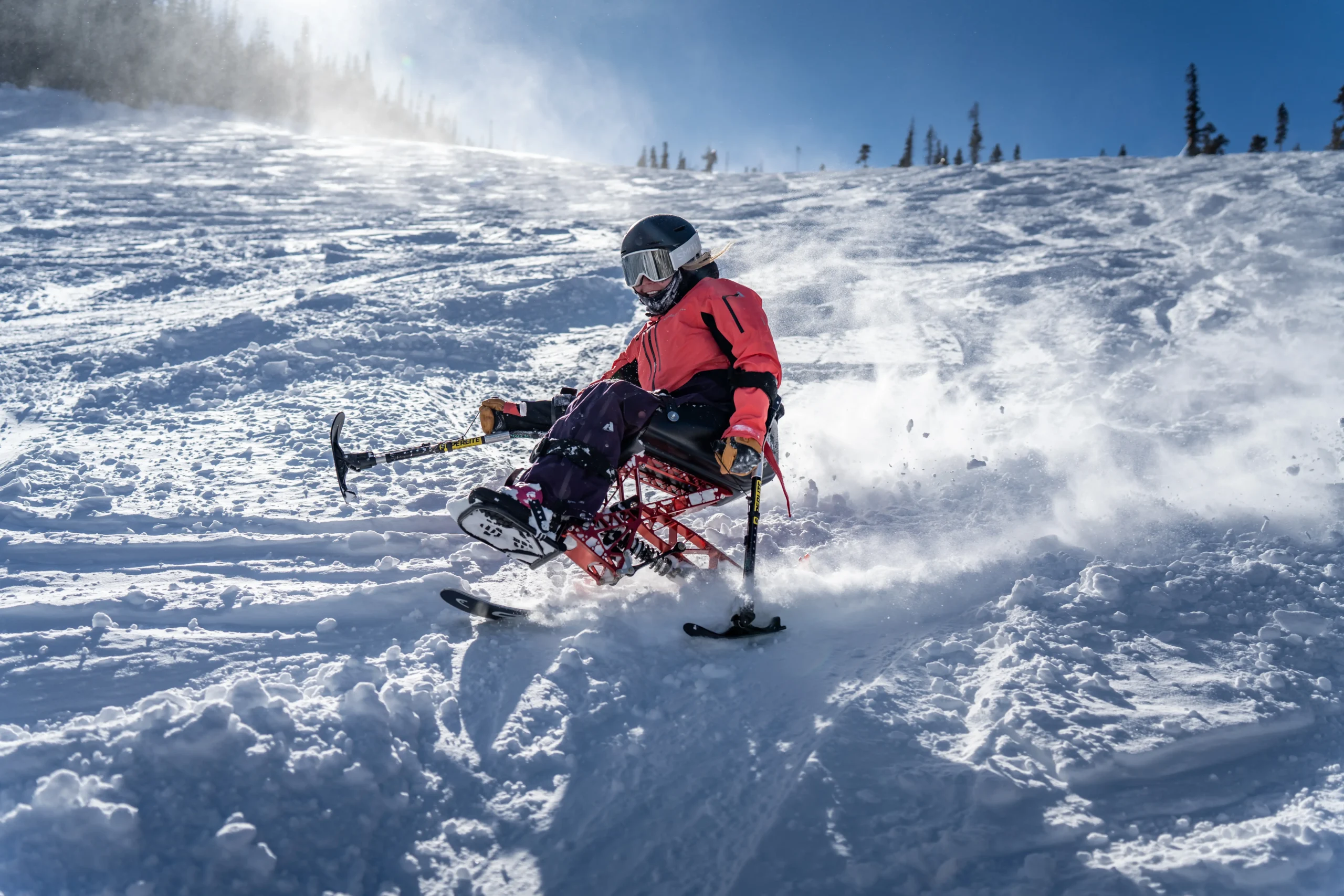 A person in a bright orange jacket maneuvers down a snowy slope on a sit ski, kicking up powder in a clear blue sky.
