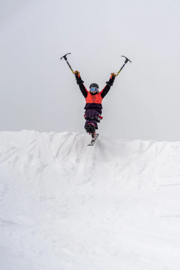 A snowboarder in an orange jacket and black gear stands triumphantly on a snowy ledge, holding ice axes overhead against a gray sky.
