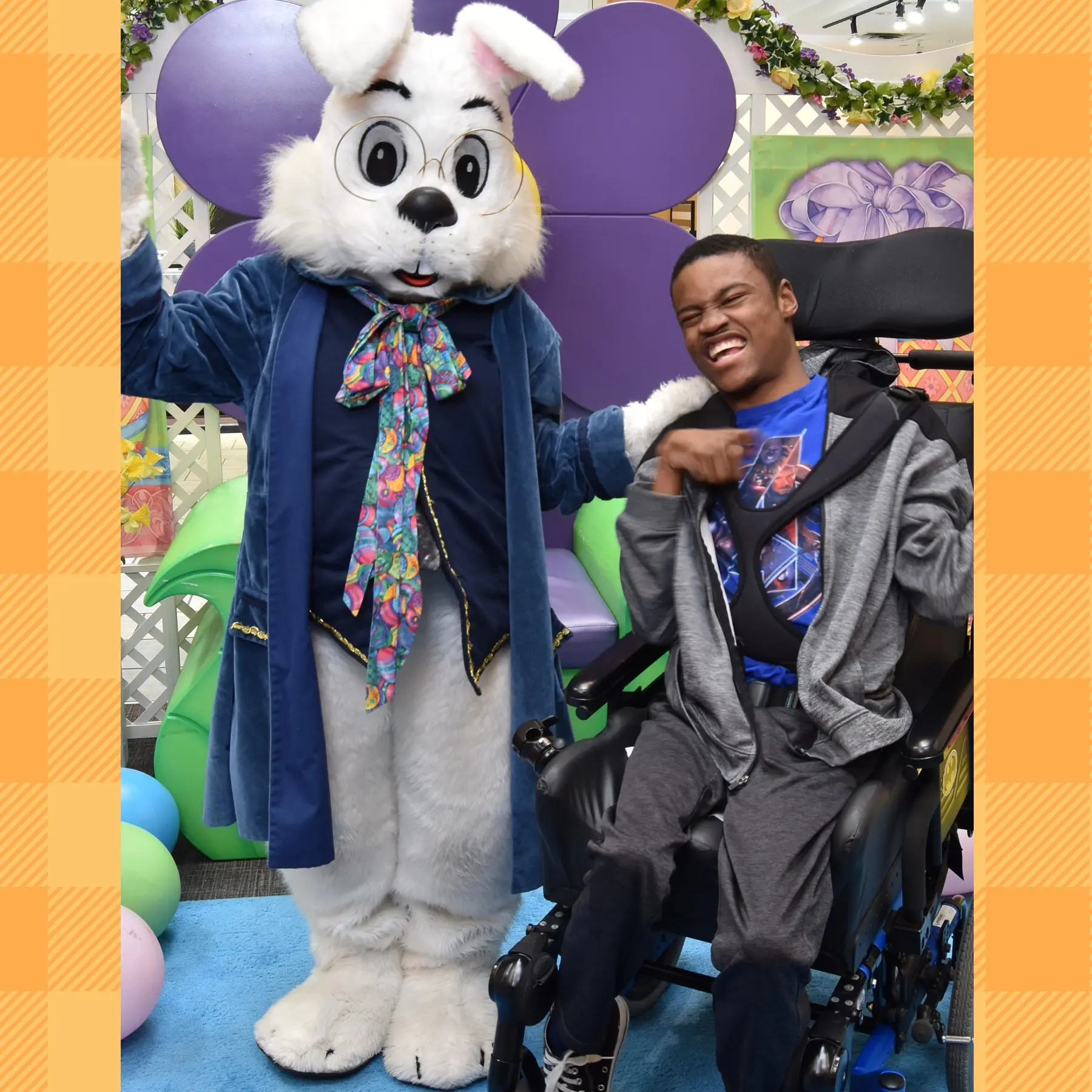 A person in a wheelchair poses with a costumed character resembling a rabbit, set against colorful balloons and decorations.