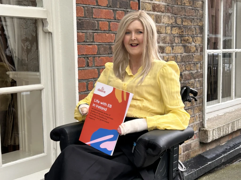 A woman in a yellow blouse sits in a wheelchair, holding a report titled "Life with EB in Ireland," against a brick wall backdrop.