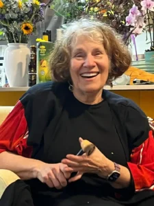A joyful elderly woman with curly, graying hair is sitting on a couch, smiling and laughing. She is wearing a black top with red sleeves and a smartwatch. In one hand, she holds a partially peeled fruit or vegetable and a knife, suggesting she is in the middle of preparing something. Behind her, a counter is adorned with colorful flower arrangements and various kitchen items, including juice and beer bottles. The atmosphere appears festive and warm.