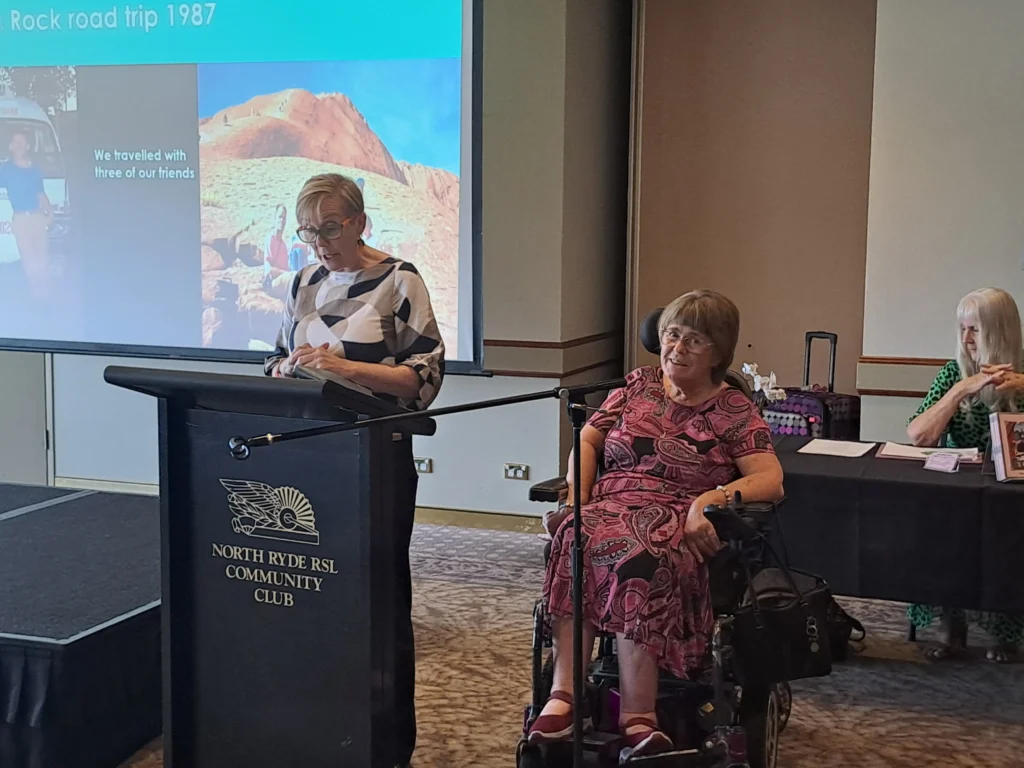 A woman speaks at a podium with a presentation behind her, while two attendees, one in a wheelchair, listen attentively in a community hall setting.