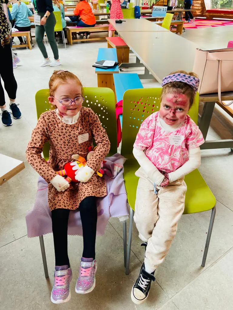 Two young girls sit on green chairs, one holding a stuffed toy, while colorful tables and people are seen in the background.