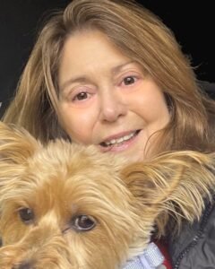 A woman with long brown hair smiles while holding a small, fluffy light-brown dog close to her face. The dog is wearing a light-colored harness and looking slightly off-camera. The background is dark, keeping the focus on the woman and her pet.
