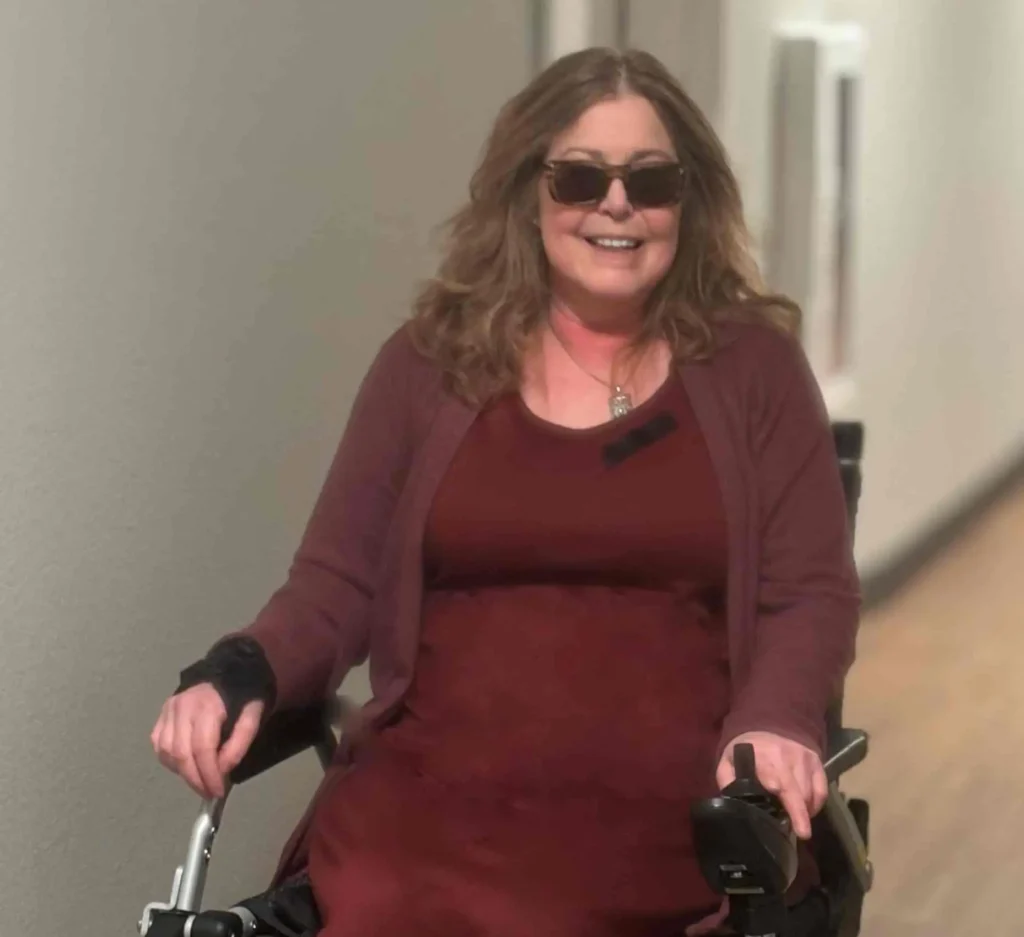 A woman with wavy, shoulder-length hair is smiling and wearing dark sunglasses while seated in a motorized wheelchair. She is dressed in a burgundy outfit with a long dress and matching cardigan. The wheelchair has a storage pouch on the side containing a water bottle. She is in a hallway with wood-patterned flooring and neutral-colored walls.