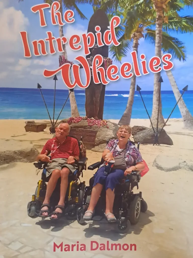 A scenic beach backdrop featuring palm trees and a stone statue, with two individuals in electric wheelchairs smiling on the sandy shore.