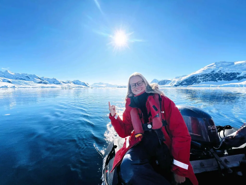 A smiling woman wearing a red parka and life jacket sits on a small boat in a calm, icy body of water surrounded by snow-covered mountains under a bright blue sky. She is pointing up toward the sun, which is shining brightly above the horizon, creating a beautiful and serene reflection on the water.