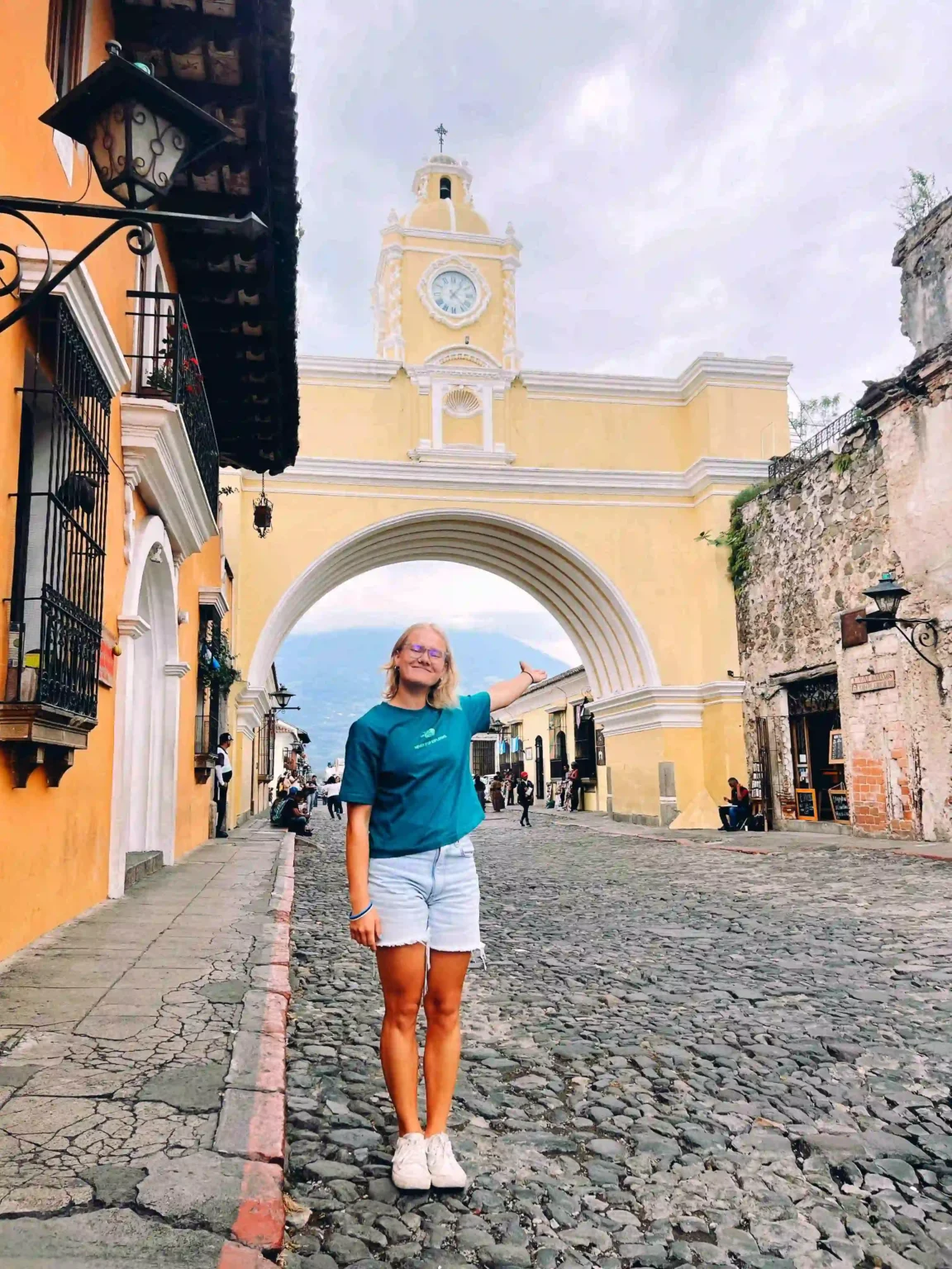 A woman in a teal shirt and denim shorts stands on a cobblestone street, smiling and pointing toward a large yellow colonial-style arch with a clock on top. The scene is in Antigua, Guatemala, with the famous Santa Catalina Arch in the background. The street is flanked by colorful buildings and a few people are walking or sitting nearby.