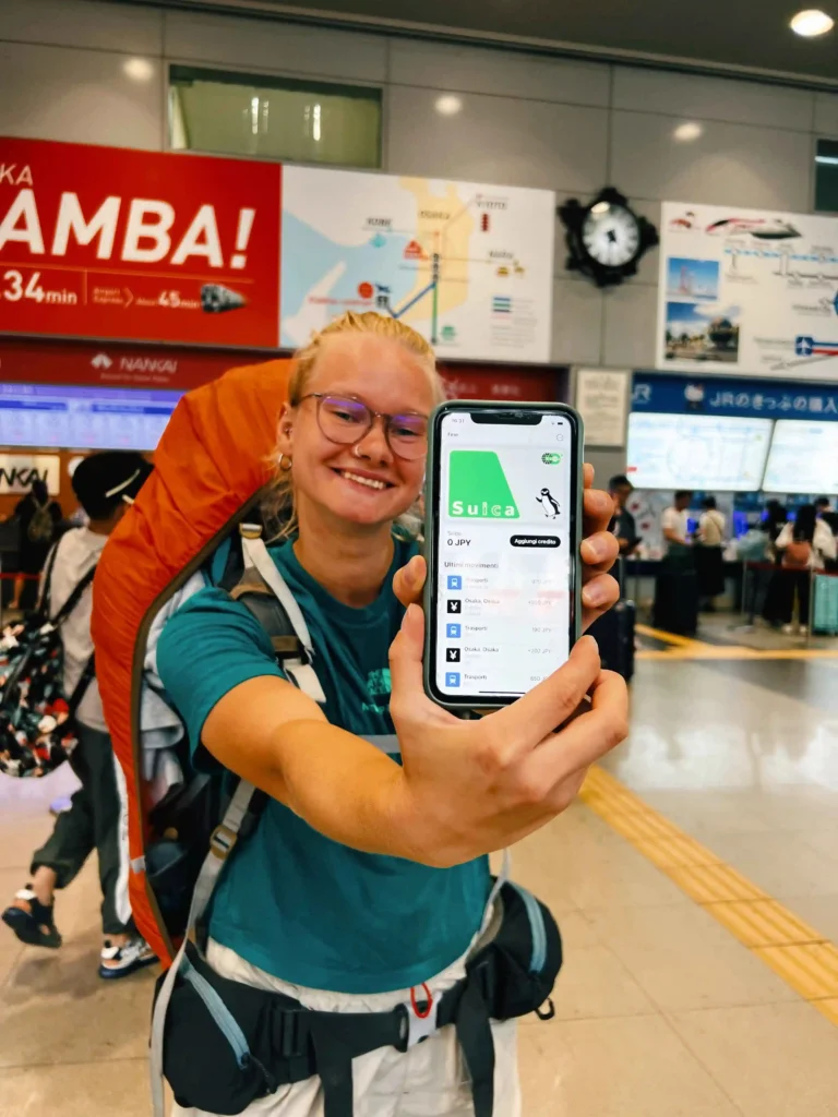 A smiling traveler with a large orange backpack is standing in a Japanese train station, holding up a smartphone displaying a digital Suica card with a 0 JPY balance. Behind them are signs in Japanese and English, a large map of train routes, and a red banner that says "NAMBA!" in bold white letters. The scene is lively, with other people in the background and a clock on the wall indicating the time.