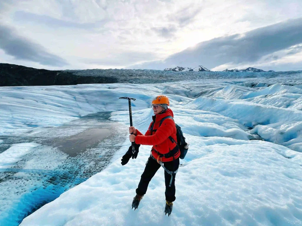 A person wearing a bright orange helmet, red jacket, black pants, and crampons stands on a vast glacier holding an ice axe. They are smiling and appear to be enjoying the icy landscape. The glacier is expansive with blue ice and crevasses, and snow-covered mountains are visible in the distance under a cloudy sky.