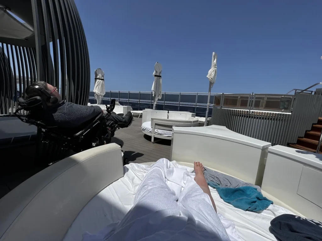 A person in a power wheelchair is relaxing under a shaded cabana on the deck of a cruise ship, wearing headphones and facing the ocean. Another person is lounging on a round, white cushioned sunbed with their legs extended, partially covered in a towel. The sky is clear and blue, and the deck features more sunbeds and covered cabanas with umbrellas closed due to the bright sun.