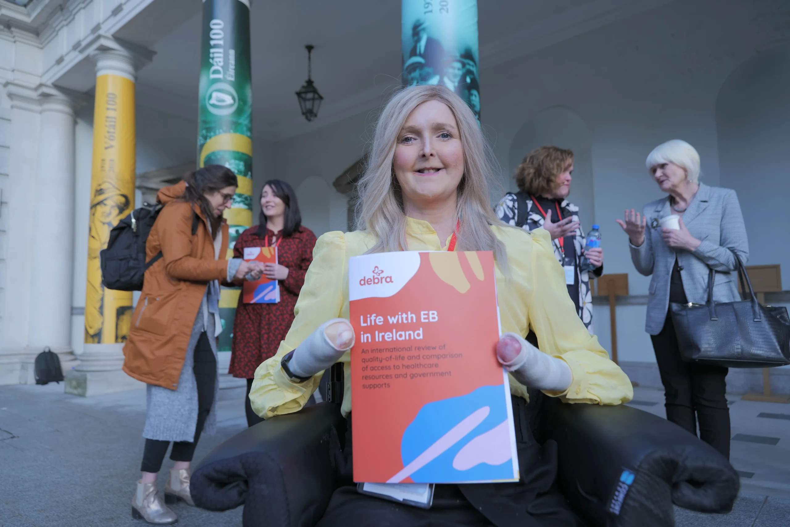 A person in a yellow blouse holds a report titled "Life with EB in Ireland" while several individuals engage in conversation behind them.