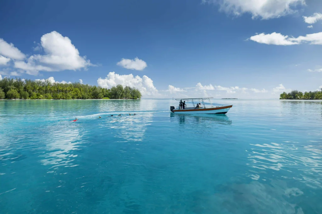 A small motorboat floats on calm, crystal-clear turquoise water near a lush, tropical island under a bright blue sky with fluffy white clouds. Several snorkelers trail behind the boat in the water, connected by a rope, enjoying the serene lagoon. Dense green trees line the distant shoreline, creating a peaceful and idyllic island scene.