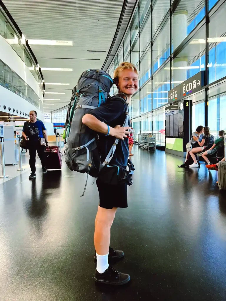 A smiling traveler wearing a large gray and blue hiking backpack stands inside a modern airport terminal. The person is dressed in black shorts, a black t-shirt, white socks, and black shoes. Other travelers and seating areas are visible in the background, along with signs pointing to various gates (EFG, BCD). The terminal has tall glass windows and a sleek, modern design.