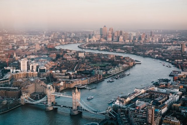 "Aerial view of London featuring the iconic Tower Bridge spanning the River Thames, with boats on the water and modern city buildings in the background under a hazy sky."