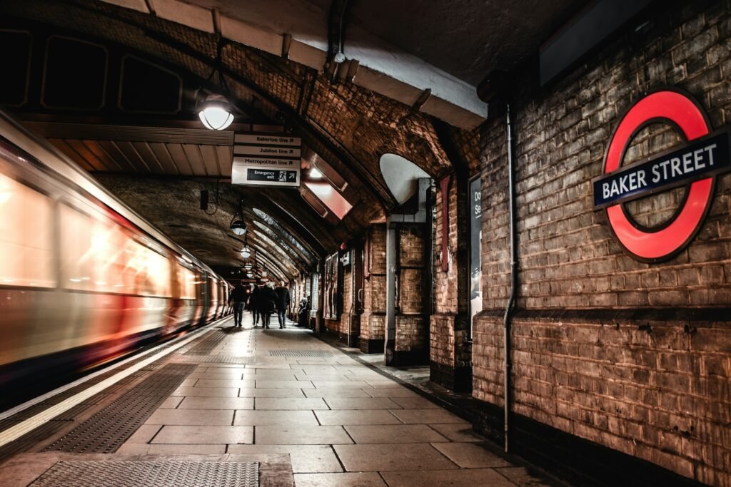 A London Underground train speeds through Baker Street station, with its iconic red roundel sign on a brick wall, arched ceilings above, and a few commuters walking along the platform."