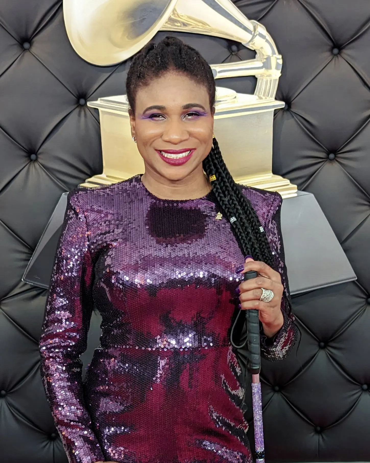 A person stands confidently in a shimmering purple sequined dress, holding a cane, with a Grammy Award statue in the background.