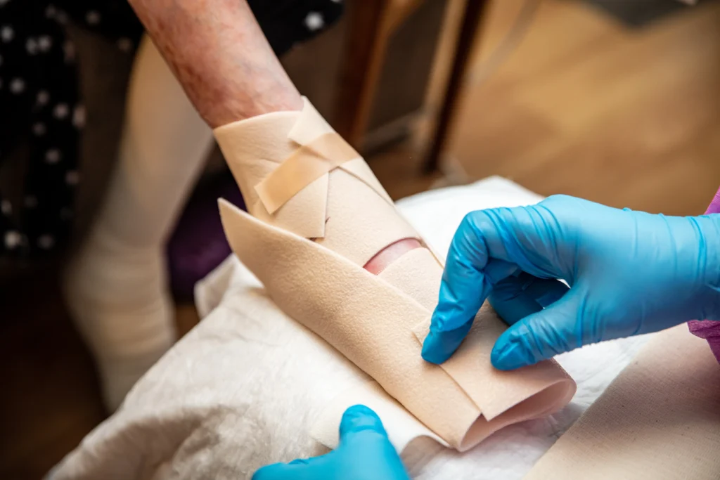 A caregiver in blue gloves wraps a bandage around an elderly person's arm, focusing on wound care and support.