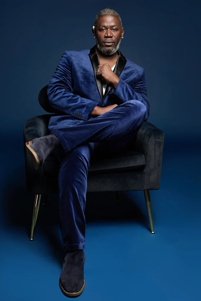 A stylish individual in a blue velvet suit sits confidently in a modern chair, against a deep blue background.