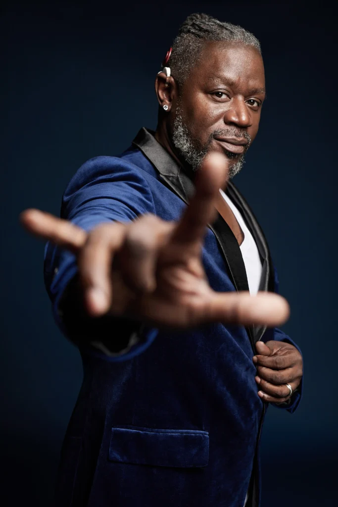 A person in a blue velvet blazer poses confidently, extending their hand forward with fingers spread, against a dark backdrop.