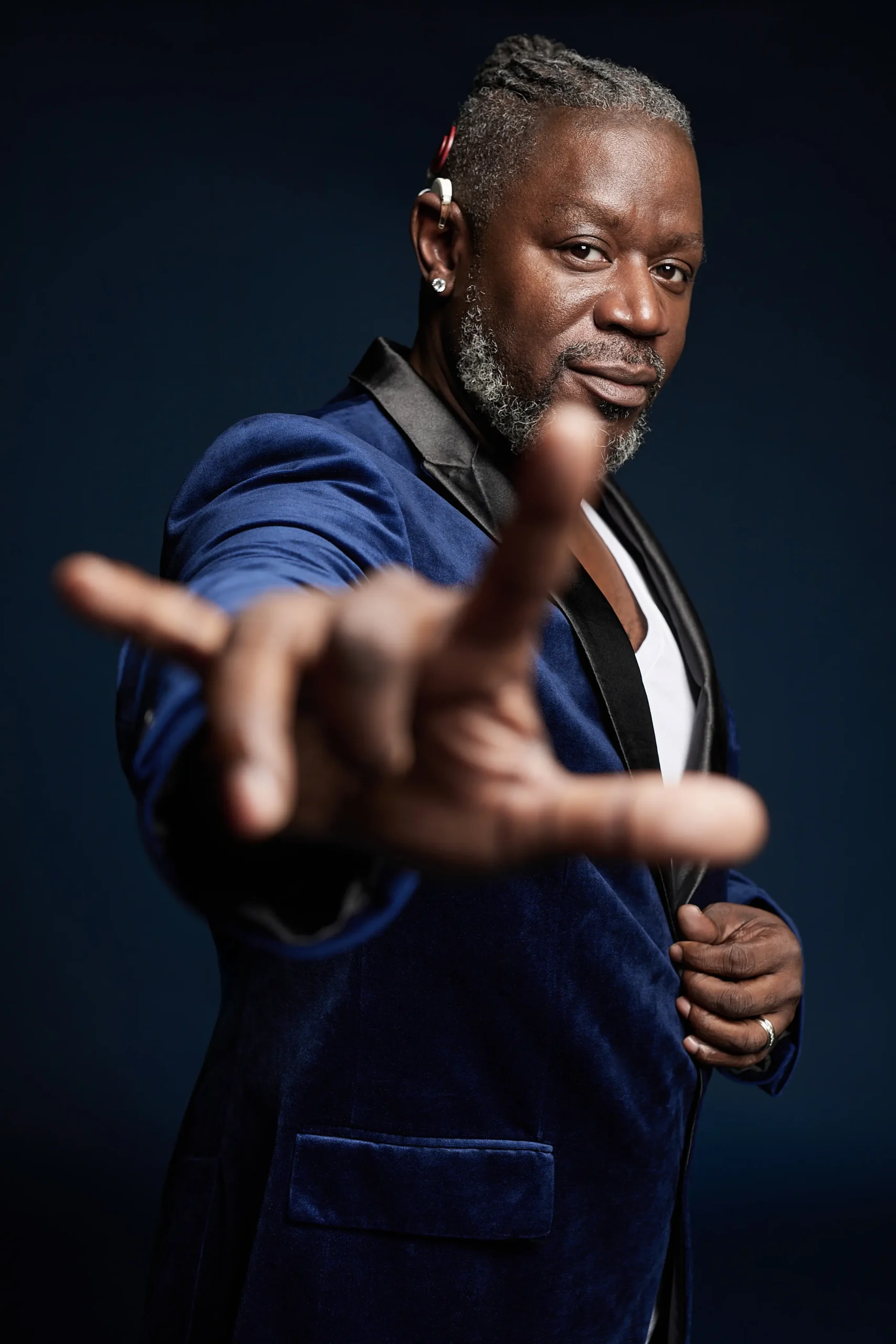 A person in a blue velvet blazer poses confidently, extending their hand forward with fingers spread, against a dark backdrop.