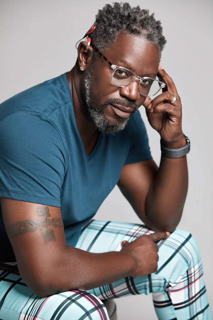 A man in a blue v-neck t-shirt and plaid pants rests his chin on his hand, showcasing a tattoo on his arm.
