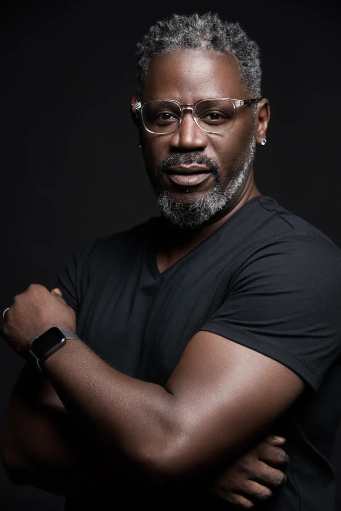 A person with short, curly hair and earrings, wearing a black v-neck shirt, crosses their arms in front of a dark background.