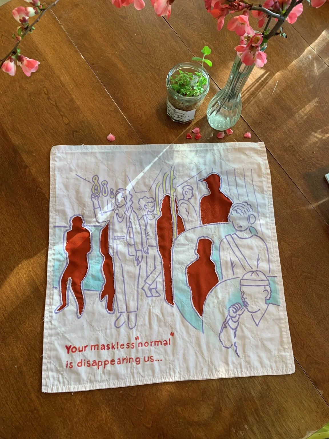 A fabric artwork lies on a wooden table next to a glass vase with pink flowers and a small plant. The artwork features outlined human figures with several filled in red, creating a layered effect. At the bottom, red text reads, “Your missed presence is disappearing us…”