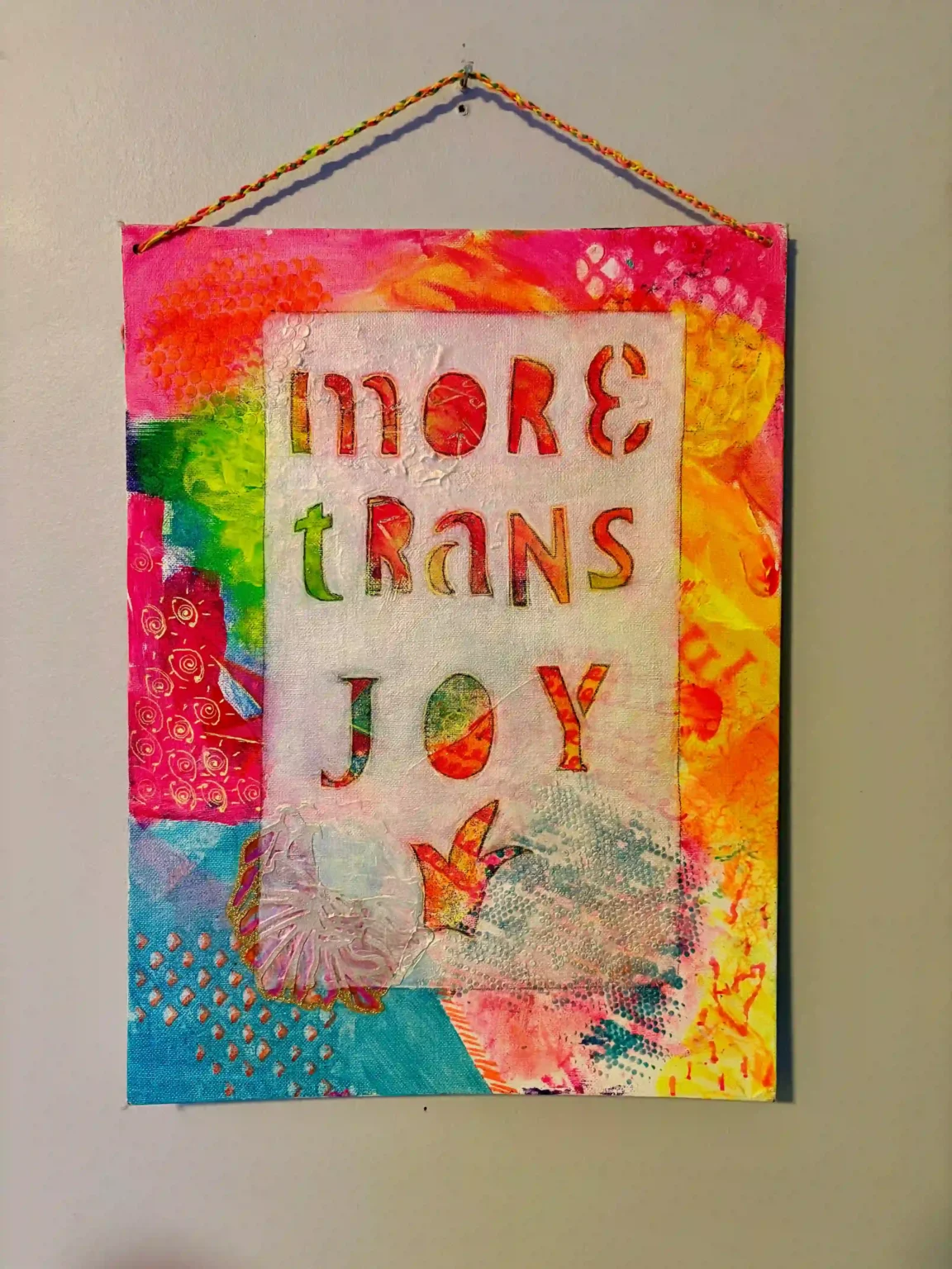 A colorful mixed-media artwork hanging on a wall with the words "MORE TRANS JOY" prominently displayed in cut-out stencil letters. The background features vibrant hues of pink, orange, green, blue, and yellow, with various textures, patterns, and abstract designs. The piece is bordered by a rainbow-colored string used for hanging.