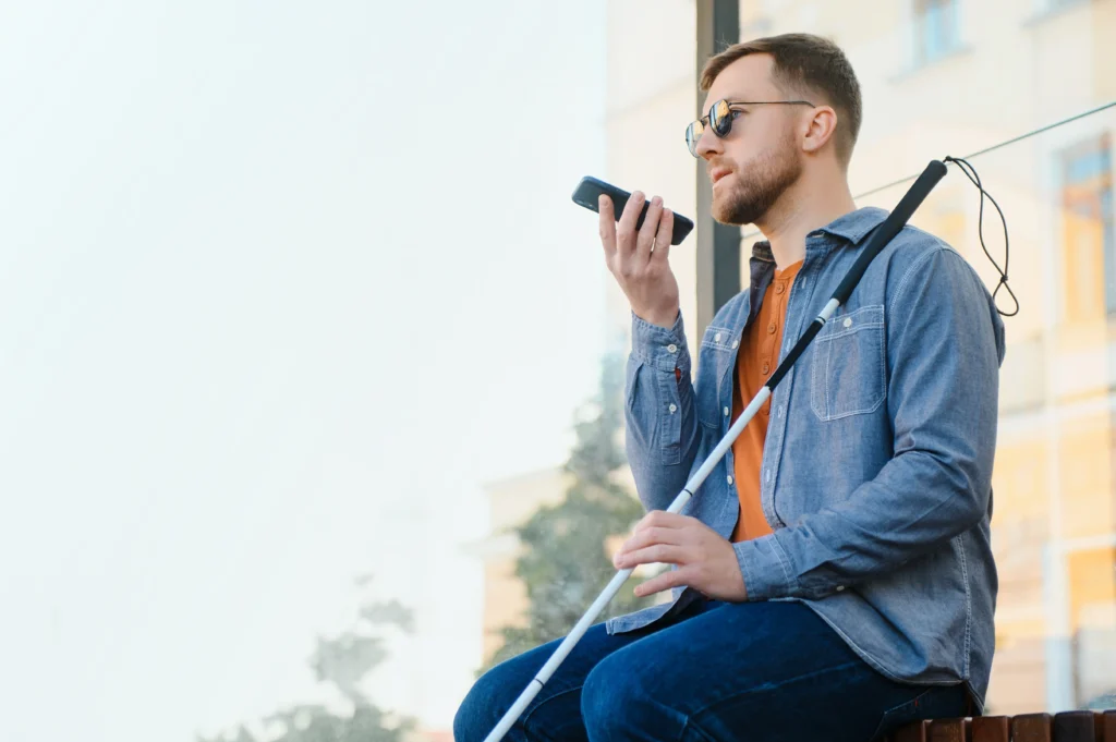 A person sitting at a bus stop using a smartphone, holding a white cane, with sunlight filtering through the glass behind.
