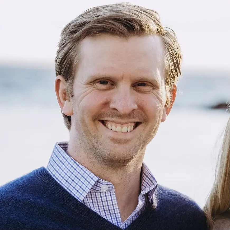 A man with light hair in a navy sweater over a collared shirt stands by the ocean, smiling beside a woman in a sunny outdoor setting.