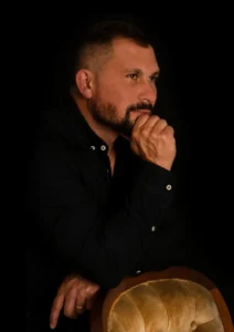 A person in a black shirt rests their chin on a fist, sitting next to a round, vintage armchair against a dark background.