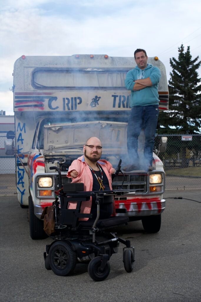 A man in a wheelchair is positioned next to a truck, with another man standing behind the vehicle. The scene takes place outdoors, featuring a clear sky and a visible road.