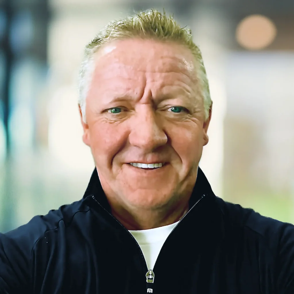A man with short, light blonde hair wears a black zip-up jacket over a white shirt, standing in a blurred indoor setting.