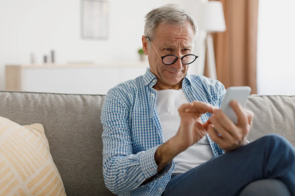 An elderly man in a plaid shirt sits on a sofa, focused on using his smartphone in a bright and cozy living room.