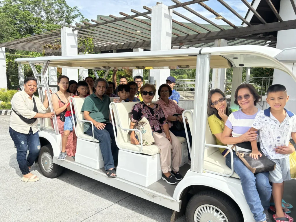 A group of diverse people sitting on a white tram, surrounded by greenery and a modern architectural backdrop, enjoying a day out.
