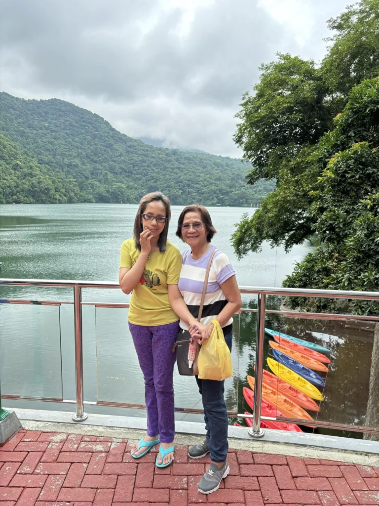 Two women pose by a serene lakeside surrounded by lush green mountains, with colorful kayaks visible in the water.