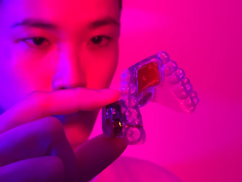 A clear dental retainer is held between fingers, illuminated in vibrant pink and purple lighting, showcasing its transparent design.