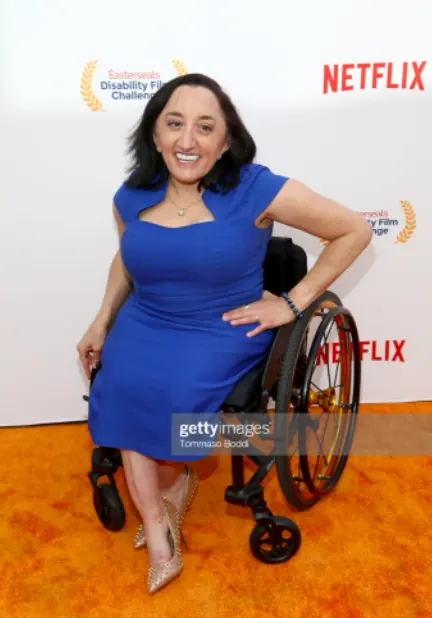 A woman in a bright blue dress sits confidently in a wheelchair, adorned with elegant heels, at a film event backdrop.