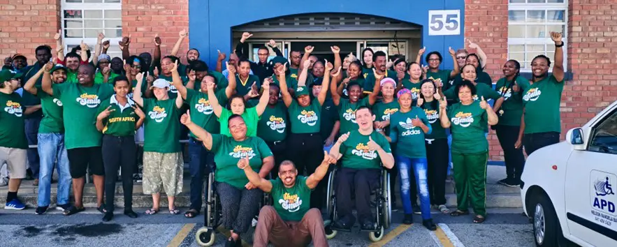 A diverse group of people in green shirts stands outside a building, raising their fists in celebration and unity.