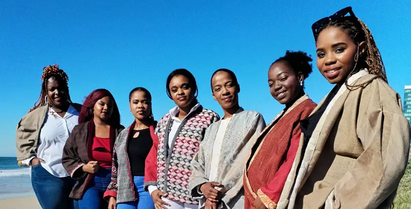 A group of seven people in colorful outfits stands together on a beach, with a clear blue sky and ocean waves in the background.