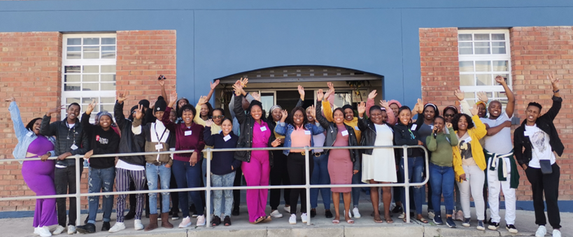 A diverse group of smiling people celebrates outdoors, posing with raised hands in front of a blue building, showcasing unity and joy.