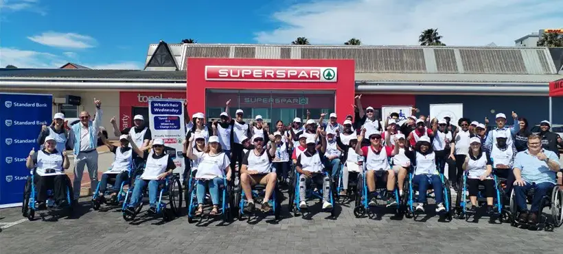 A group of wheelchair users in matching t-shirts smiles and poses in front of a SUPERSPAR store on a sunny day.