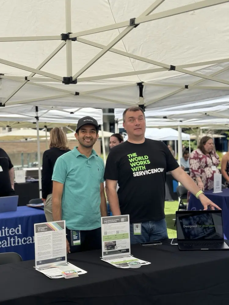 Two individuals stand at a booth under a white tent, showcasing brochures and a laptop featuring a digital display, surrounded by tables.