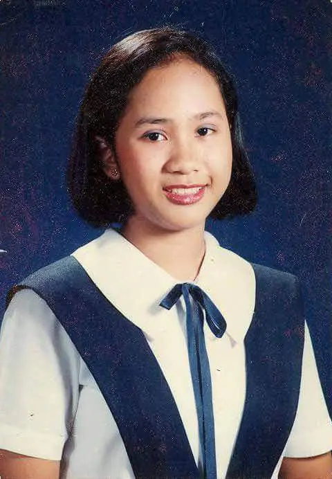 A young person in a school uniform with a white shirt and navy blue dress, standing against a dark blue background.