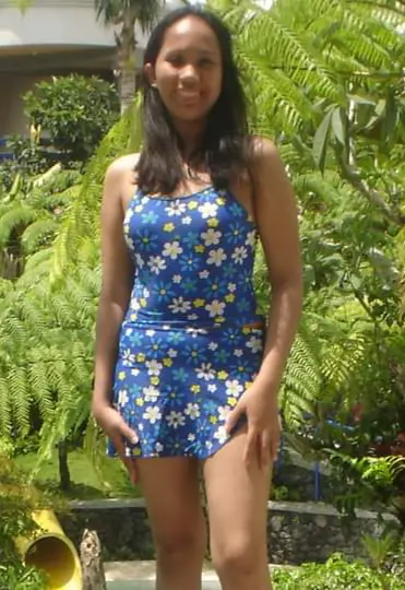 A young woman stands in a sunlit garden, wearing a blue floral dress, surrounded by lush green plants and tropical foliage.
